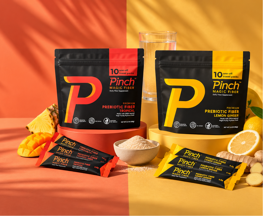 Two stand-up pouches of Pinch Magic Fibers prebiotic fiber, one in Tropical Mango flavor and one in Lemon Ginger flavor, displayed with their single-serve packets, a glass of water, and citrus slices on a split orange and yellow background.
