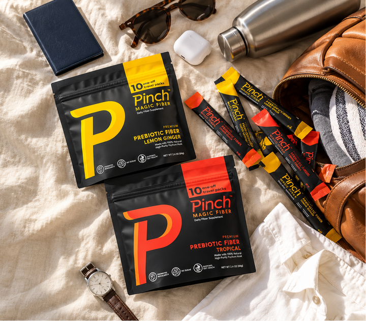 Two pouches of Pinch Magic Free Prebiotic Free Fiber are displayed in a styled flat-lay arrangement on a beige fabric background, surrounded by lifestyle props including sunglasses, a watch, a water bottle, a wallet, and a brown leather bag.