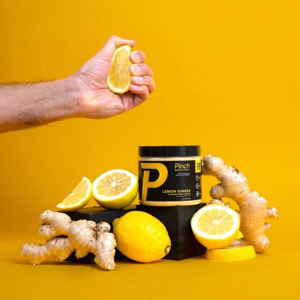 A hand squeezes a lemon over a jar of Pinch lemon ginger powder surrounded by fresh lemons and ginger.
